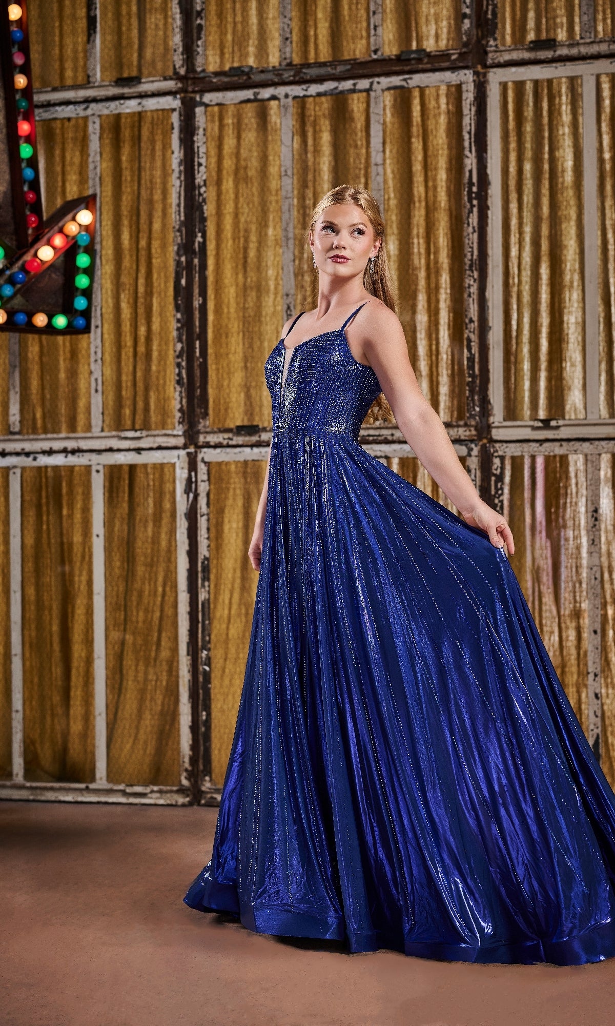 A woman stands indoors in front of gold curtains and a large lit arrow sign, wearing the Tiffany 16189 long royal blue sleeveless A-line prom dress with a full skirt.